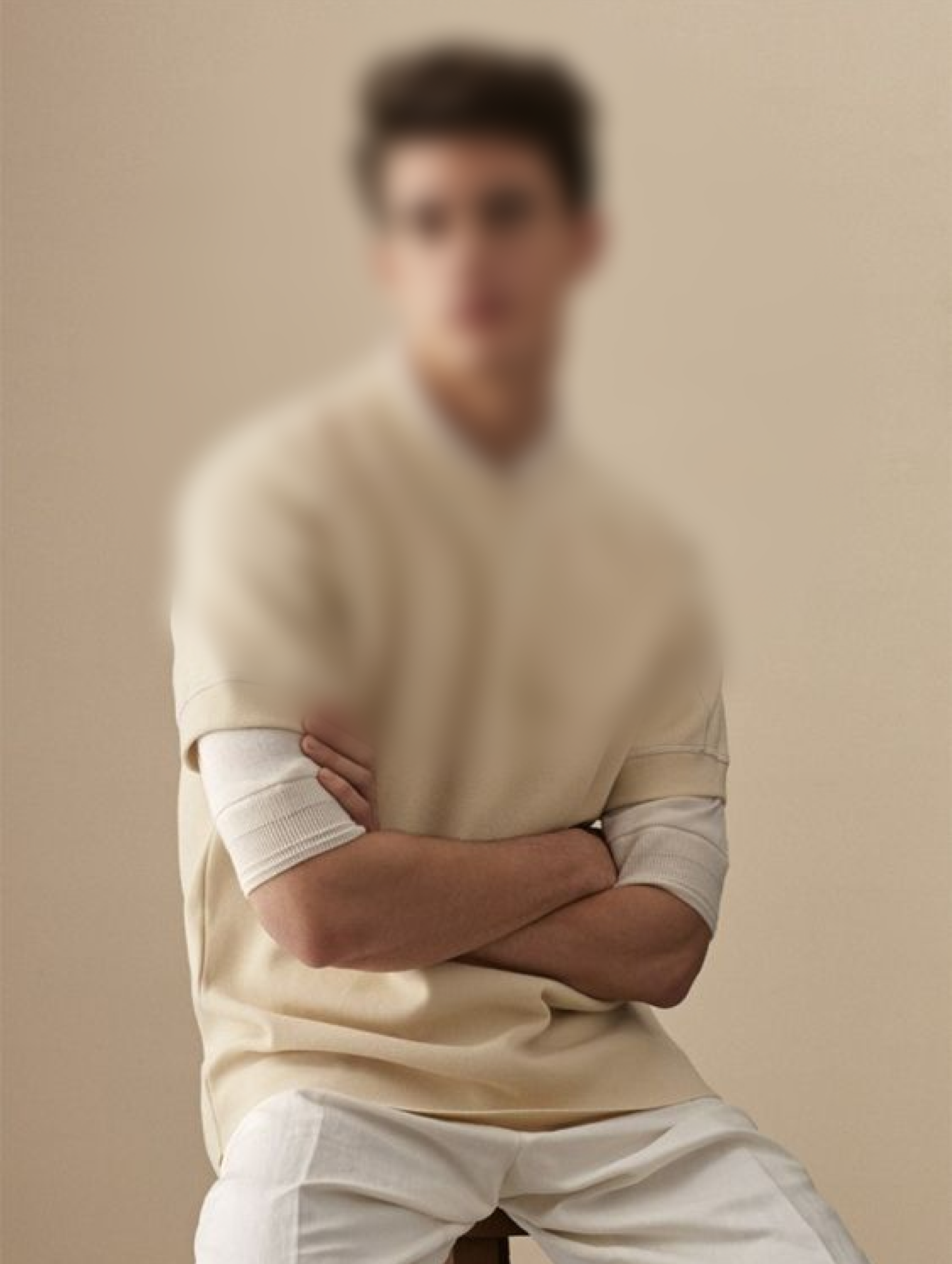 A male model sitting on a stool in white pants and a off white shirt.