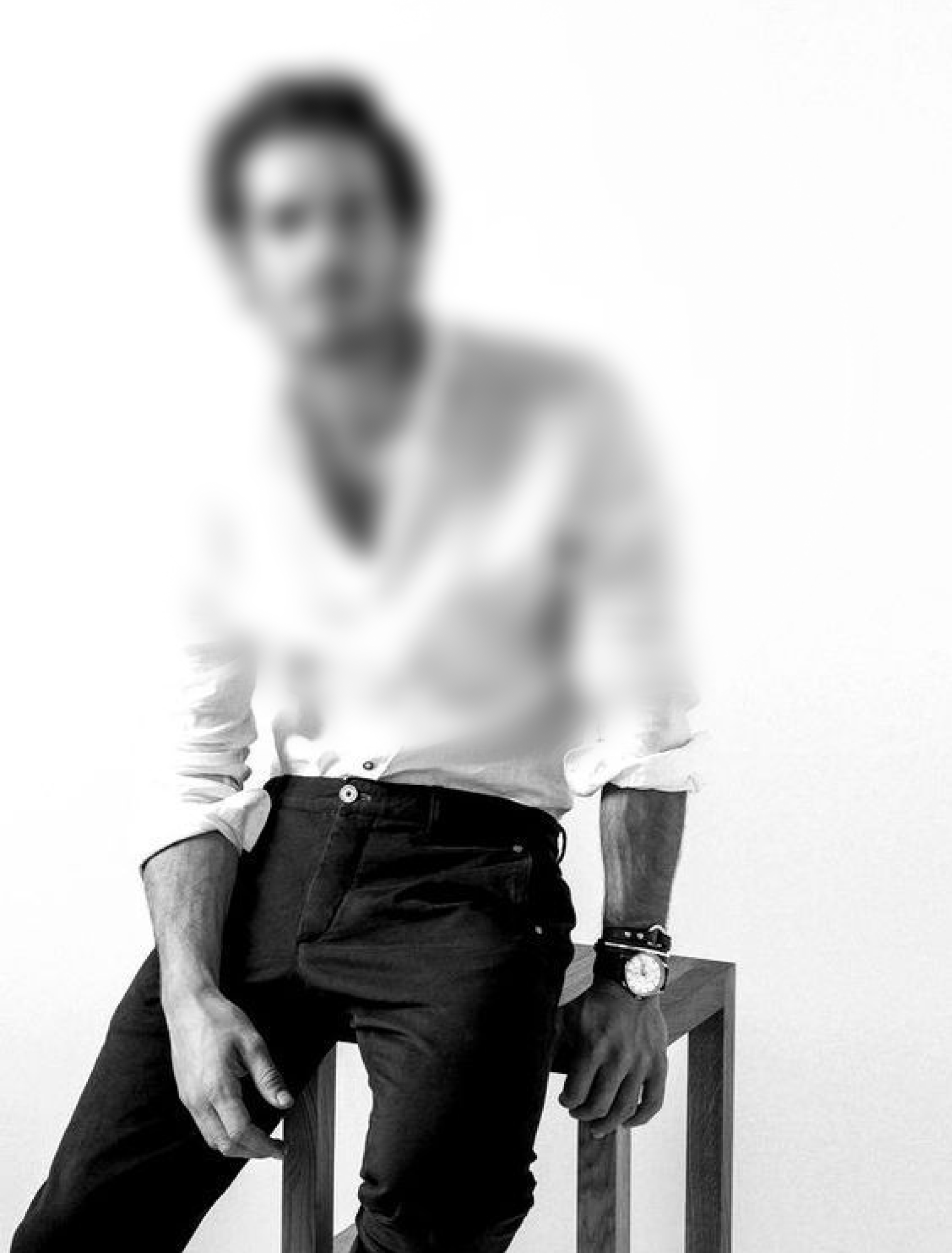 A male model sitting on a stool, wearing a white shirt and black pants.