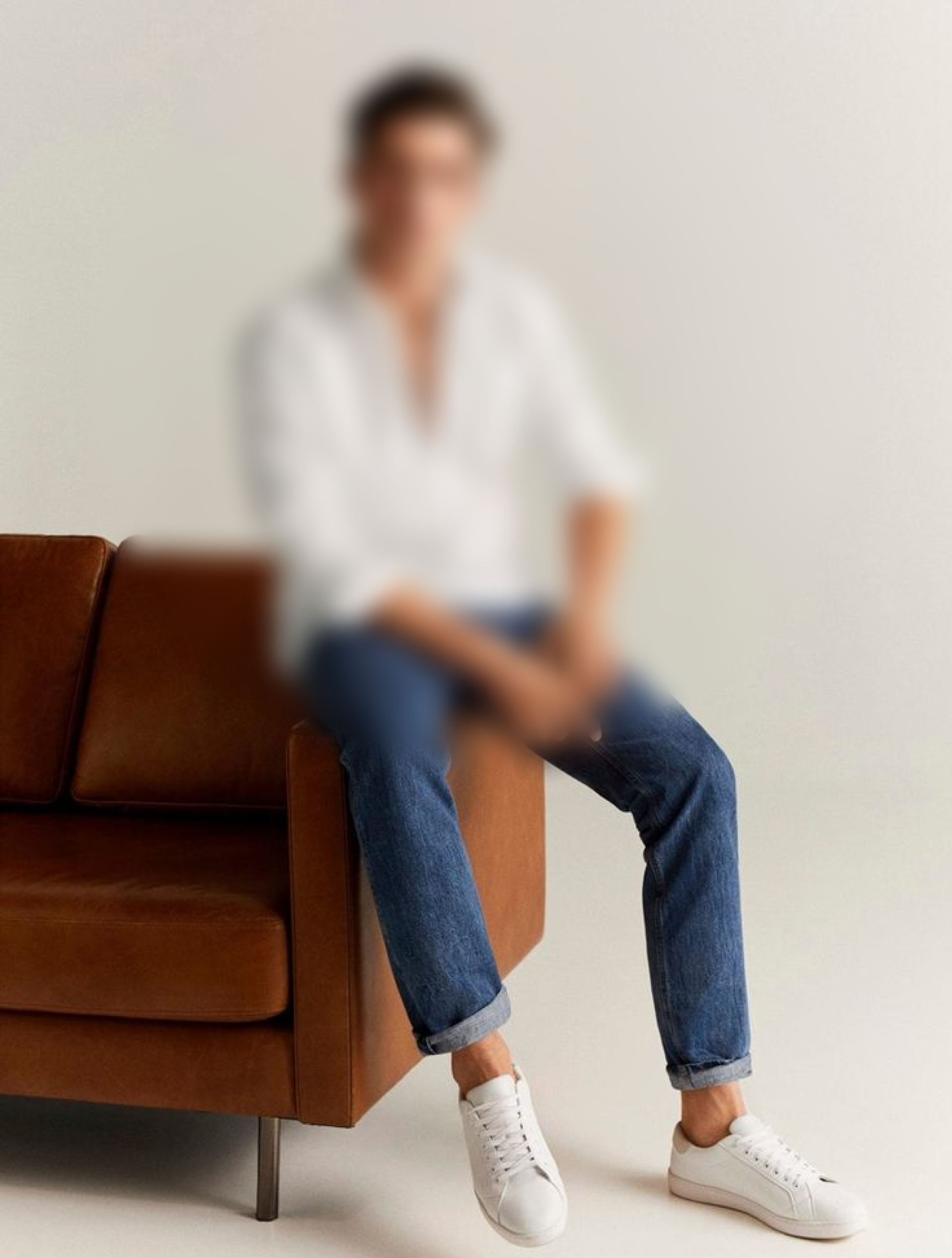 A young male model sitting on a brown couch waring blue jeans and white shirt and sneakers